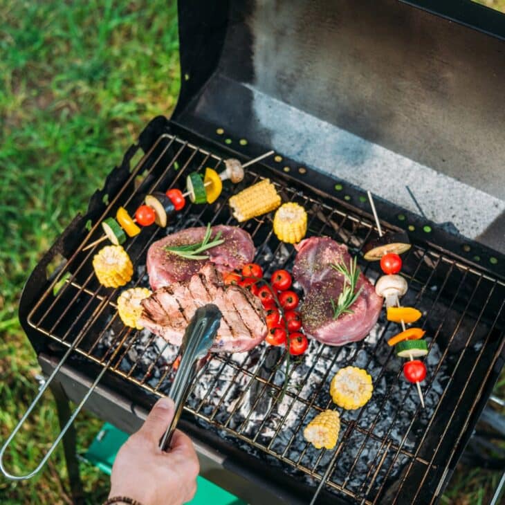 A person uses tongs to grill assorted vegetables, corn cobs, and two large pieces of meat seasoned with rosemary on an outdoor barbecue grill set on grass.