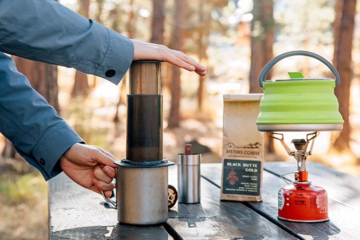 Megan using an aeropress to make a cup of coffee. A camp stove and kettle are in frame on the table.