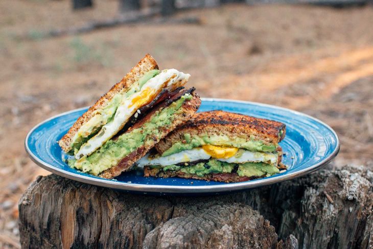 Breakfast sandwich with avocado, egg, and bacon on a blue camping plate placed on a log