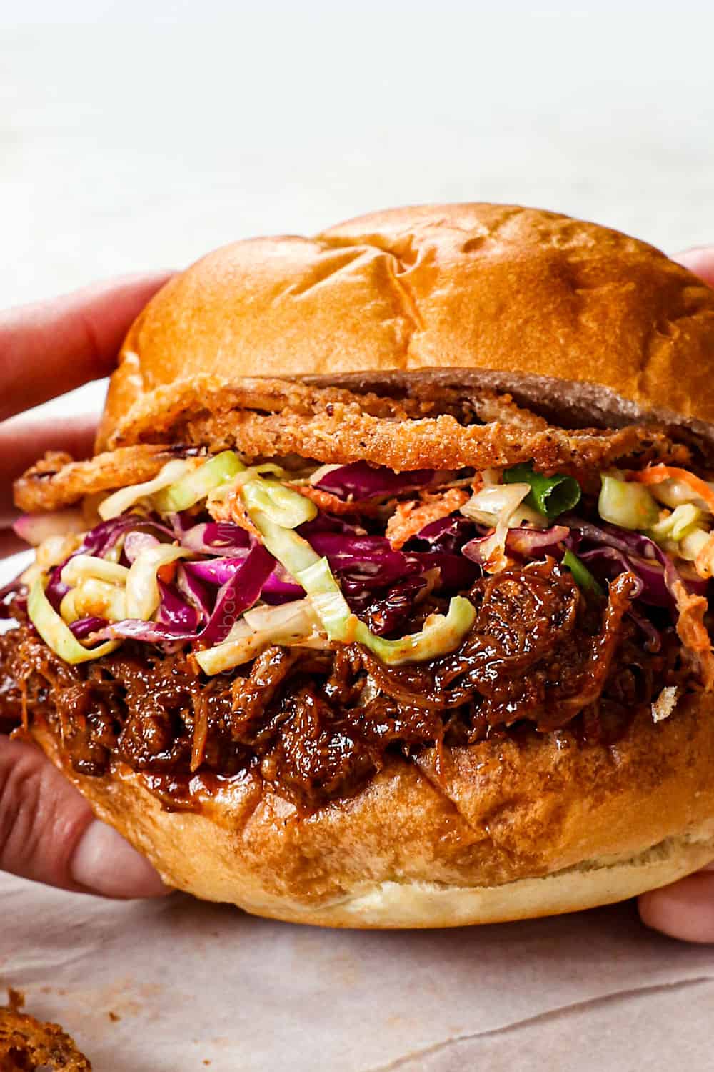 A hand holds a barbecue pulled pork sandwich with crispy onions and coleslaw nestled in a golden bun.