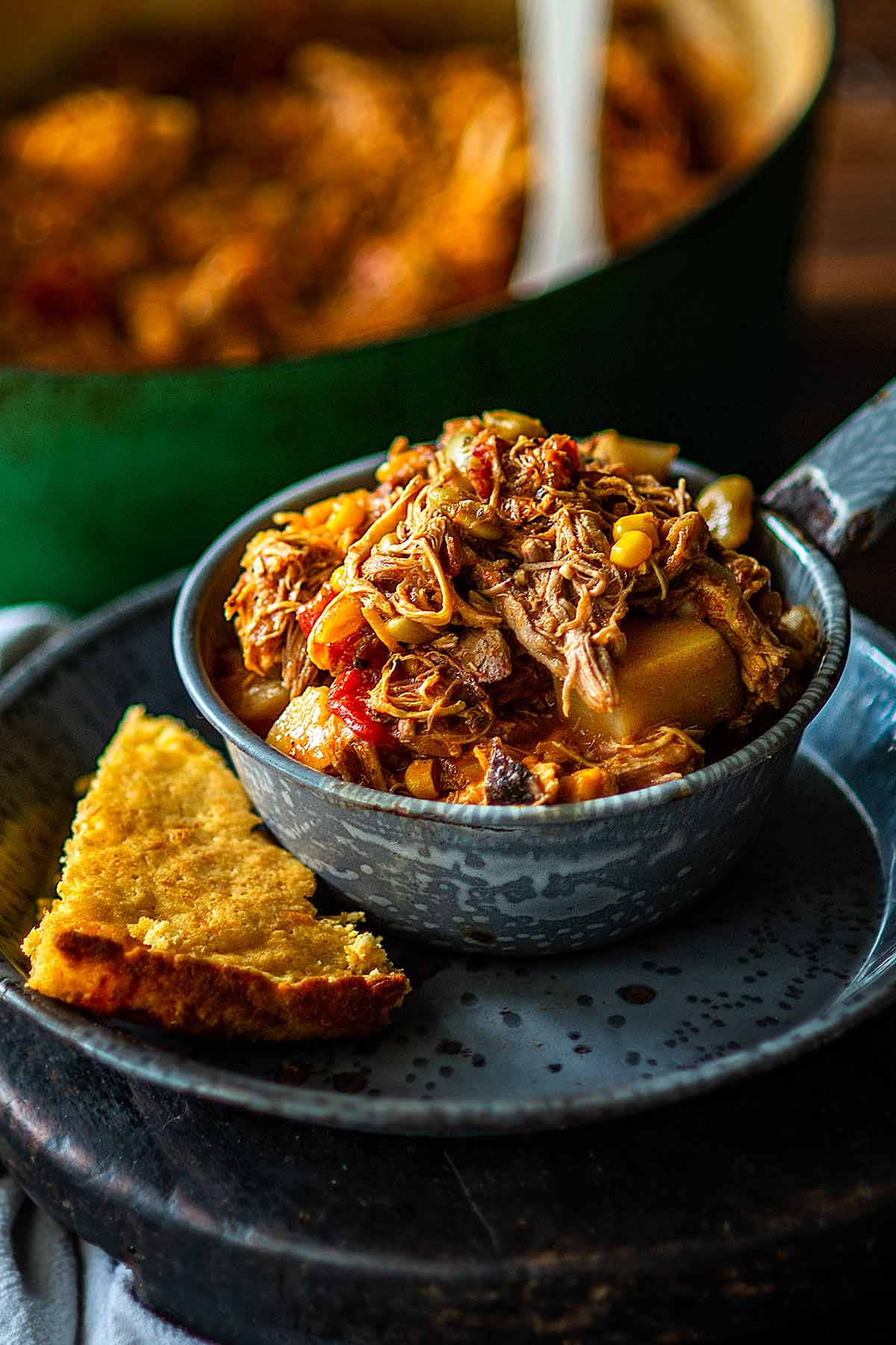 A bowl filled with shredded meat, corn, and tomatoes sits next to slices of golden-brown cornbread on a speckled plate, with a pot in the background.