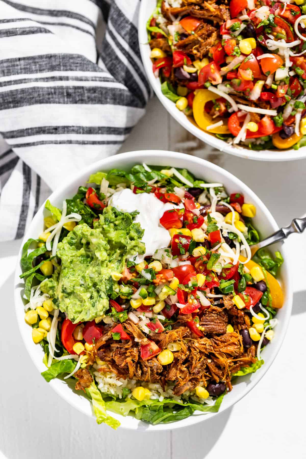 Two vibrant bowls hold a pulled pork salad garnished with guacamole, sour cream, corn, tomatoes and mixed greens on a white surface beside a striped napkin.