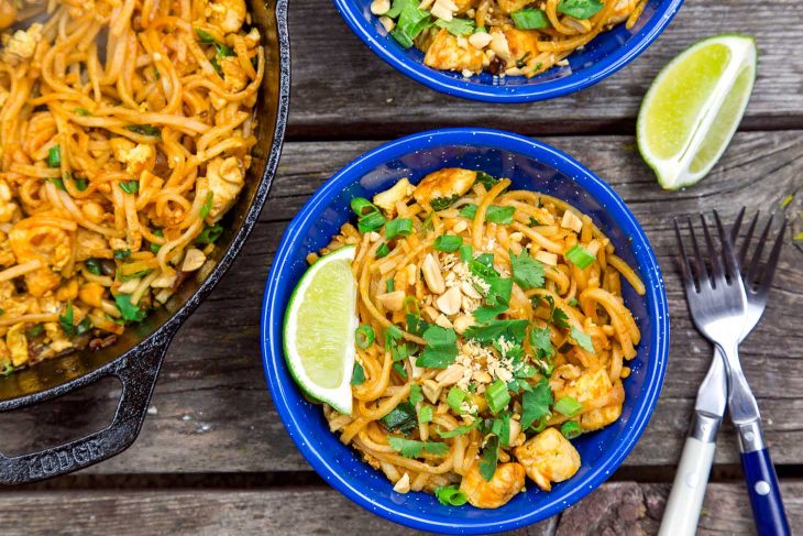 Two bowls of Pad thai and a wedge of lime next to a skillet on a wooden surface