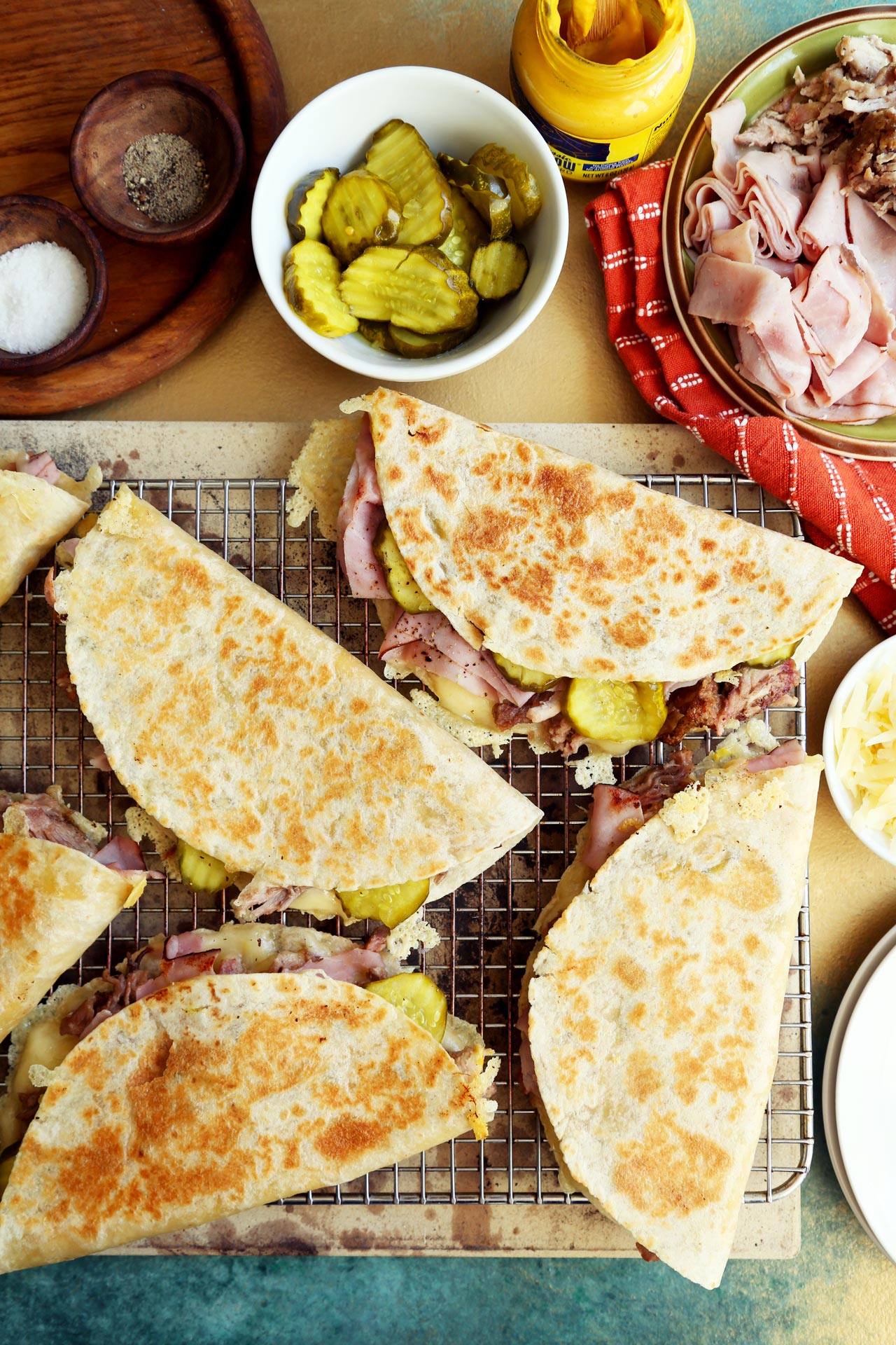 Cuban sandwiches are displayed on a wire rack, encircled by pickles, mustard, sliced meats, cheese and seasoning.