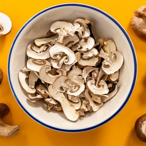 A bowl of sliced mushrooms surrounded by whole mushrooms on a yellow background.