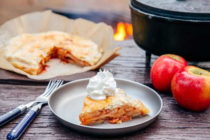 Slice of apple pie on a plate with a Dutch oven and campfire in the background