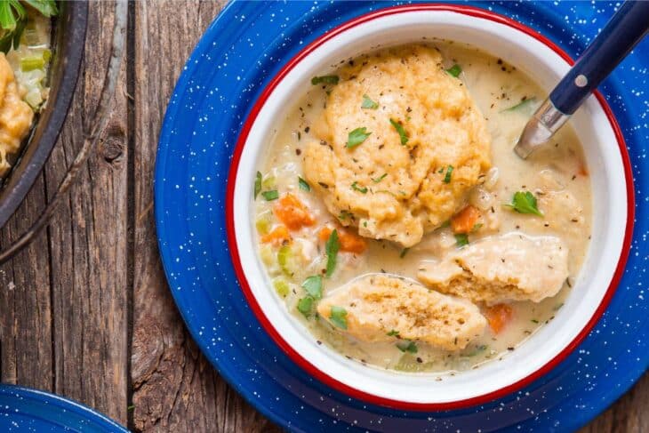 A bowl of creamy chicken and dumplings soup, garnished with herbs, sits on a blue speckled plate on a rustic wooden table with a spoon resting inside.