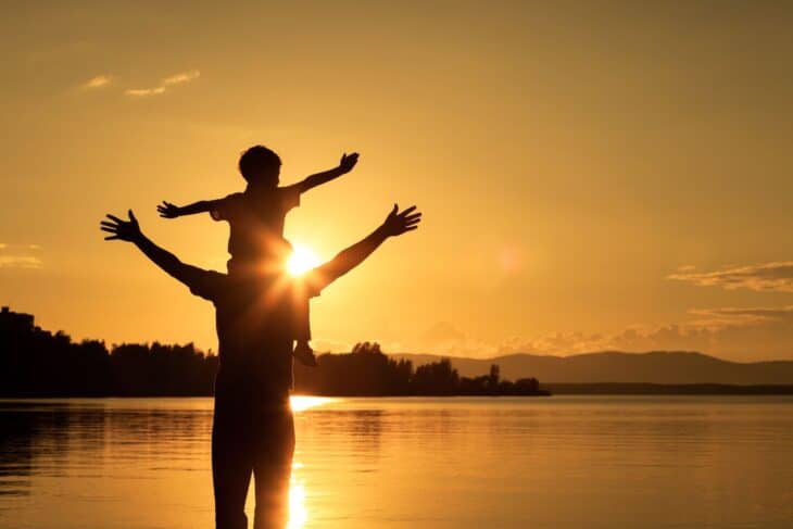 Silhouette of a child sitting on a parents shoulder with a lake in the background.