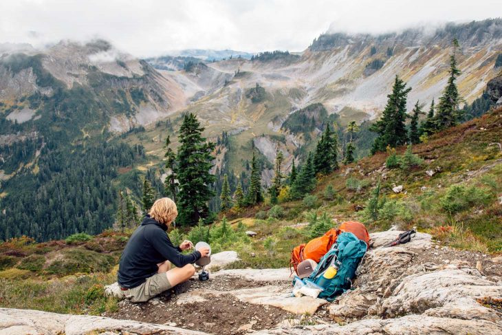 Man cooking a backpacking meal with mountains in the background.