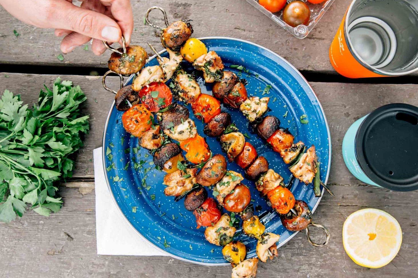 Four chicken and vegetable kebabs on a blue camping plate Michael is picking one up