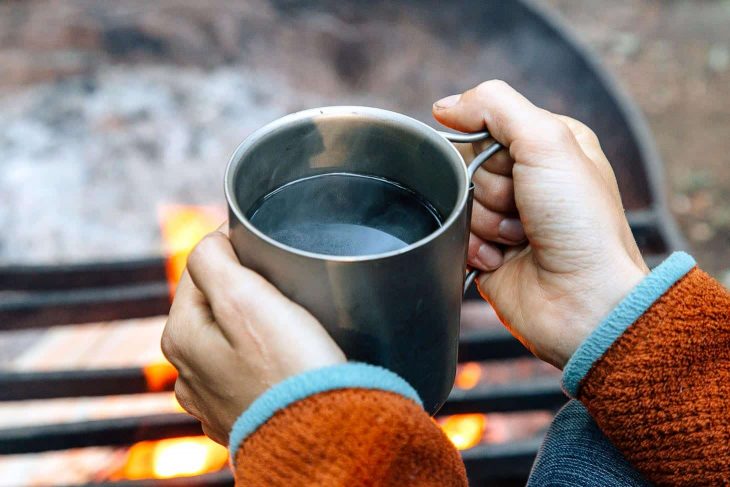 Megan holding a cup of coffee with a campfire in the background