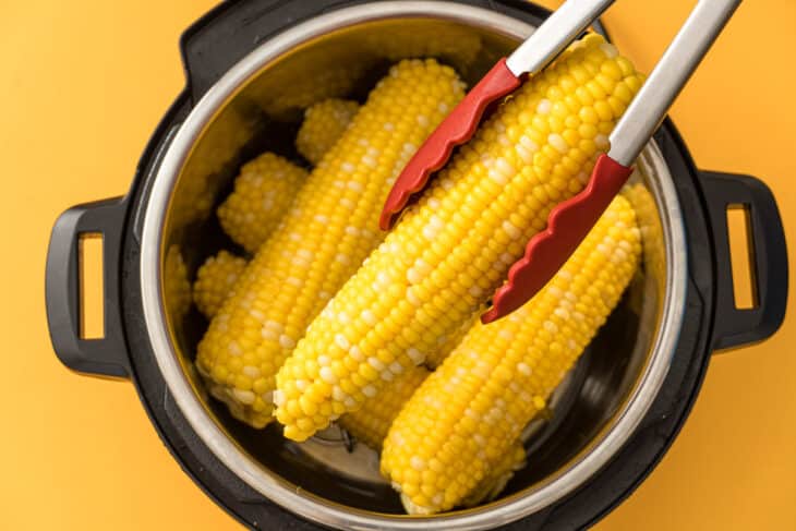 A pot filled with freshly cooked corn on the cob, with yellow and white kernels visible, and a pair of tongs with red tips lifting one cob out of the pot against a yellow background.