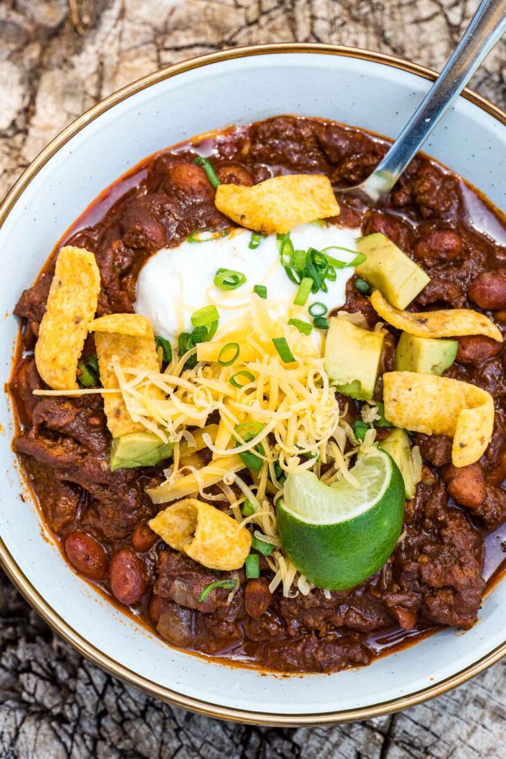 A bowl of chili topped with shredded cheese, diced avocado, sour cream, corn chips, and chopped green onions sits on a rustic wooden surface with a lime wedge on the side and a spoon tucked into the bowl.