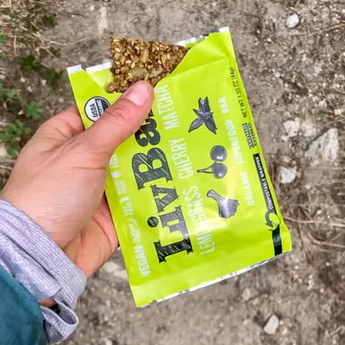 A person holds an open green snack pouch labeled "LivBar" with a small portion of a cherry matcha bar visible, standing on a dirt trail.