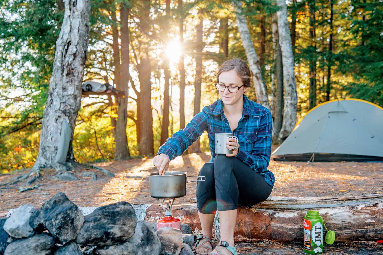 Megan is sitting on a rock next to a campfire stirring a pot of food