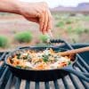 Michael sprinkling cheese over a skillet of pasta with a desert scene in the background