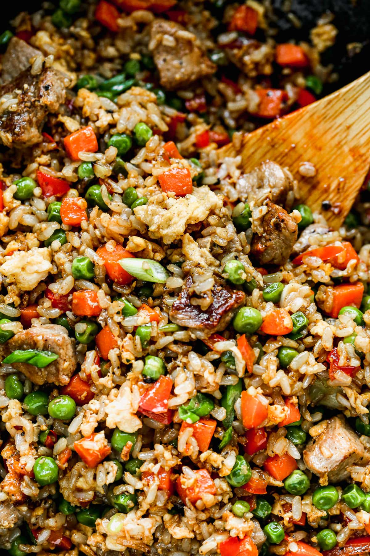 A colorful skillet contains a mixture of fried rice, green peas, diced carrots, eggs, and chunks of meat with a wooden spoon.