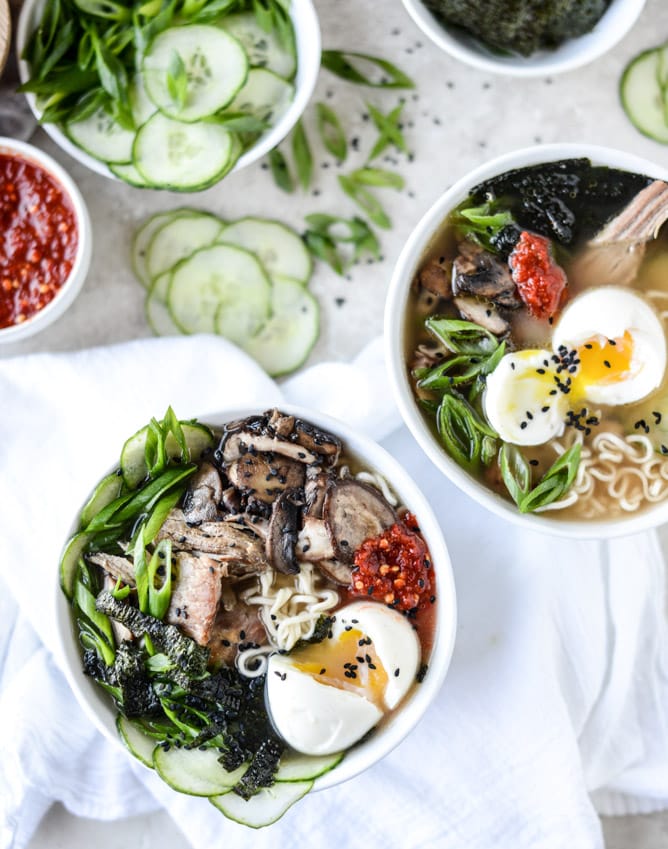 Two bowls are filled with ramen, eggs, mushrooms, cucumbers, green onions and red sauce; alongside these rest a heap of cucumber slices and a separate bowl of broth.