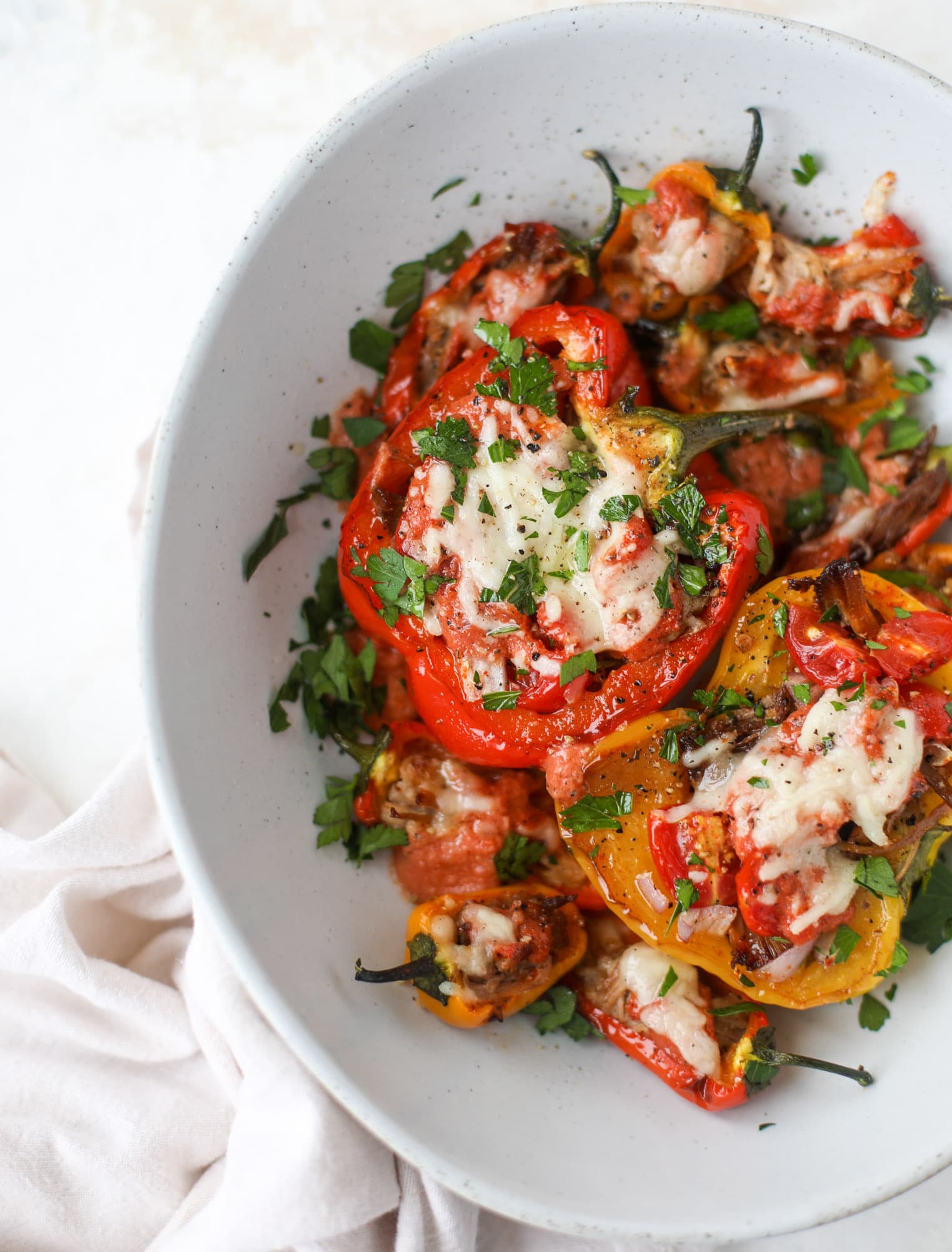 A bowl holds baked bell peppers filled with cheese and herbs, their vibrant colors topped with melted cheese and chopped parsley, all set on a white rustic table.