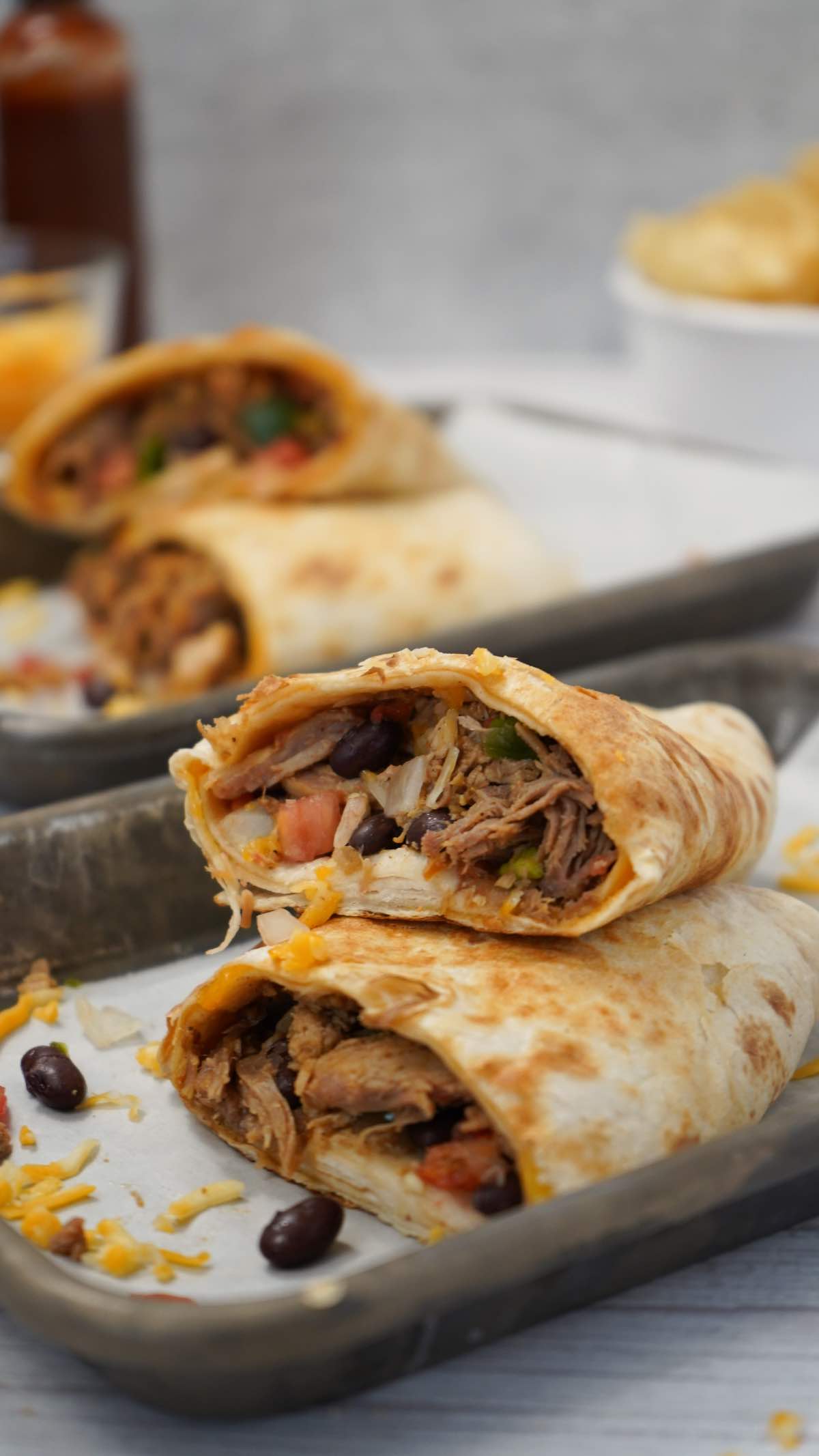 A halved burrito, stuffed with beef, beans, and vegetables, sits on a metal tray in frame's foreground while additional burritos and a chip bowl occupy the background.