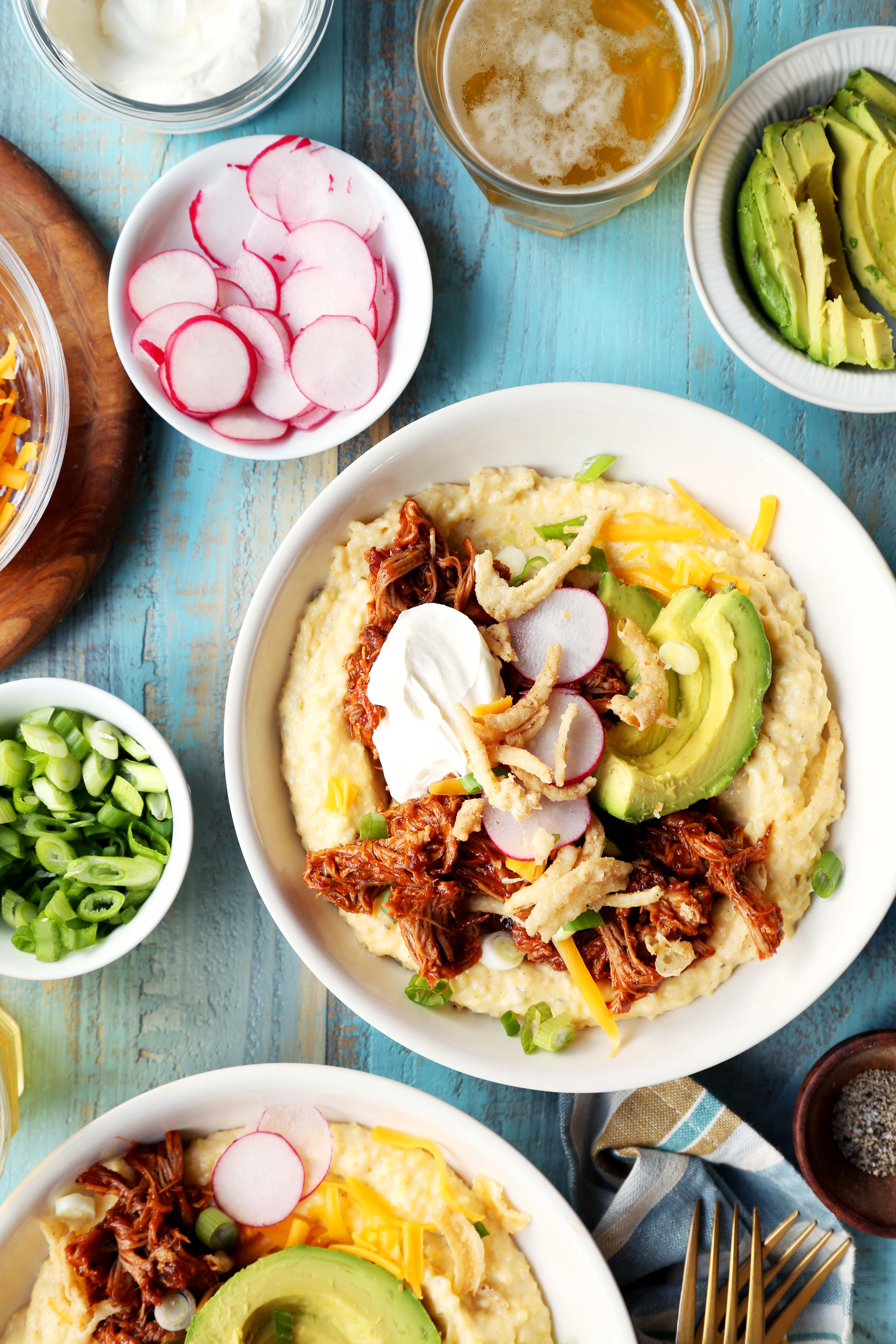 A plate, holding pulled pork, avocado slices, radishes, and sour cream atop mashed potatoes, sits on a blue wooden table surrounded by small bowls of ingredients.