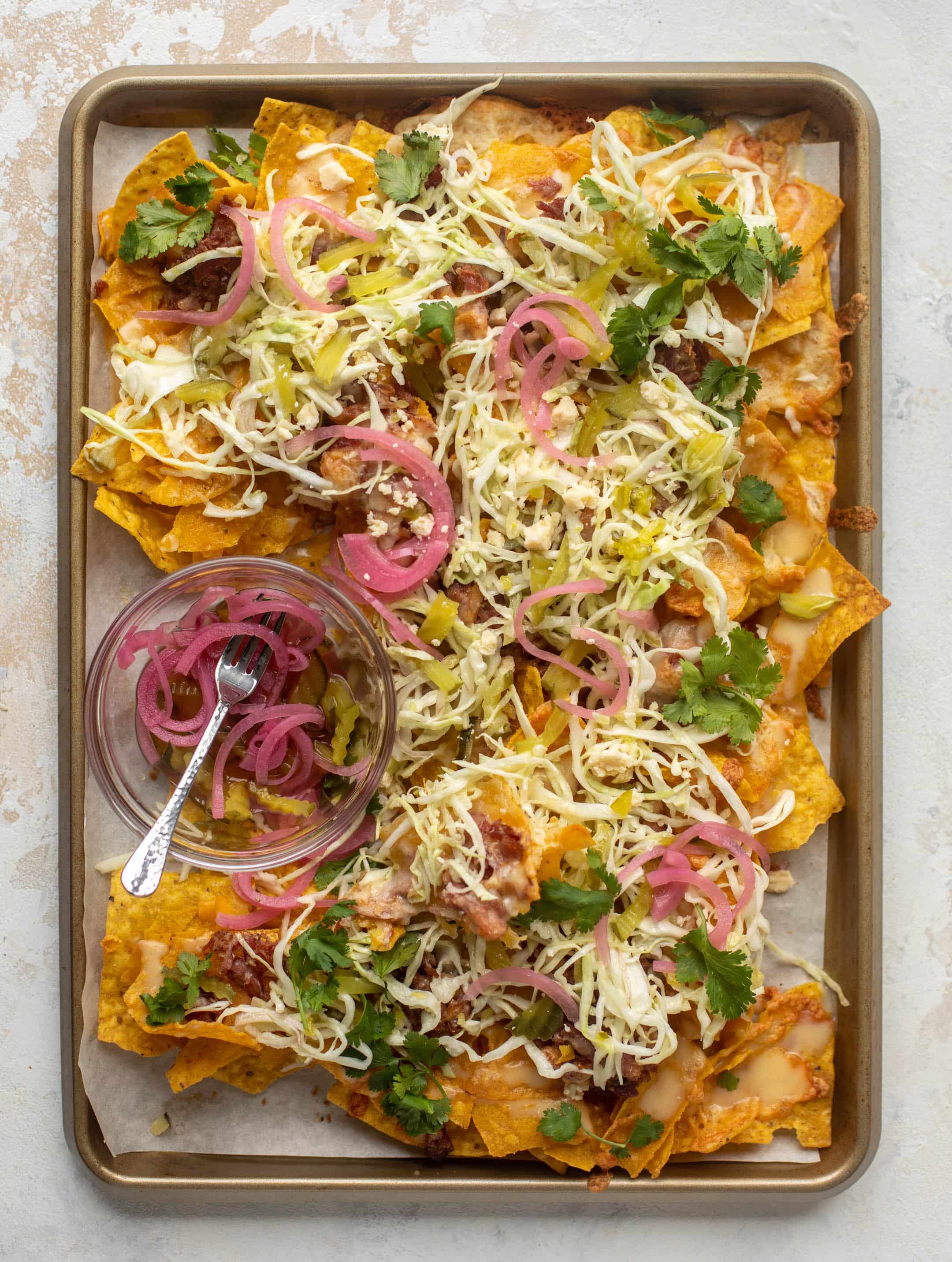 A tray is filled with nachos, covered in melted cheese, adorned with jalapeños, pink pickled onions, cilantro and shredded lettuce.