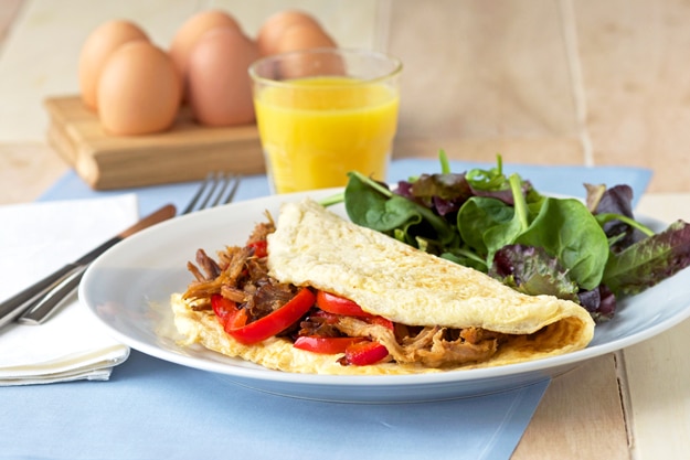 A folded omelet with red peppers on a white plate, next to a salad, eggs, and a glass of orange juice on a wooden table.