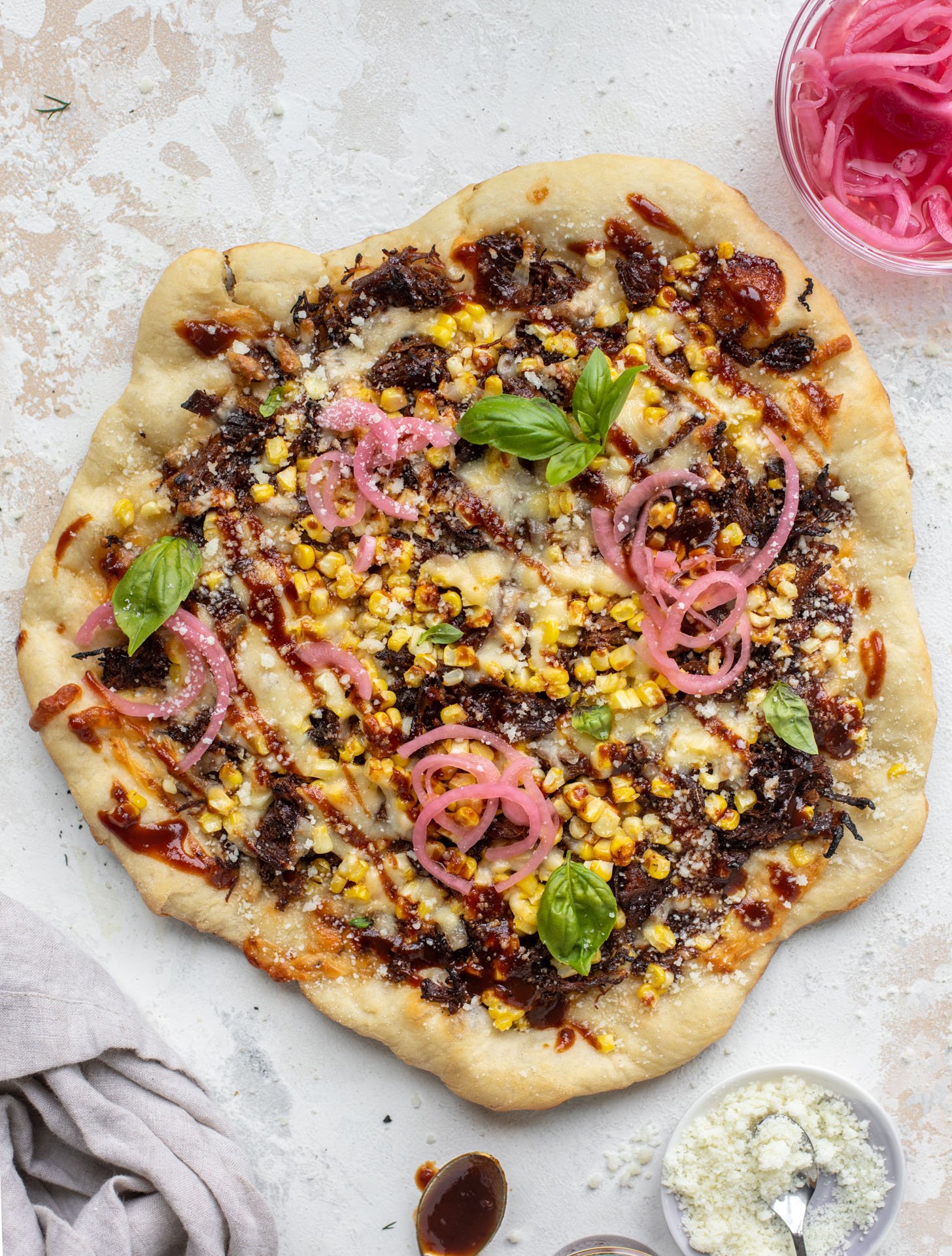 A pizza, topped with corn, caramelized onions, pink pickled onions, barbecue sauce and basil leaves rests on a textured countertop next to a dish of crumbled cheese and pickled onions.