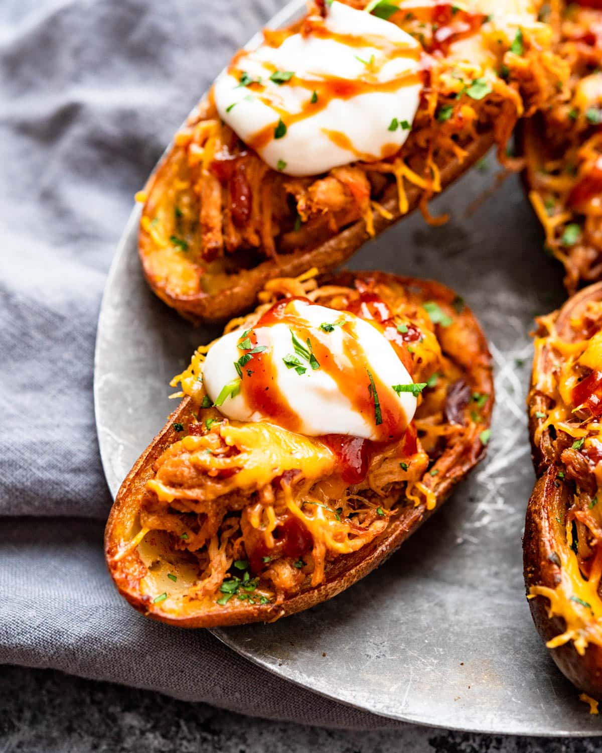 A plate of potato skins is garnished with melted cheese, bacon bits, a dollop of sour cream, and chopped parsley on a textured gray surface.