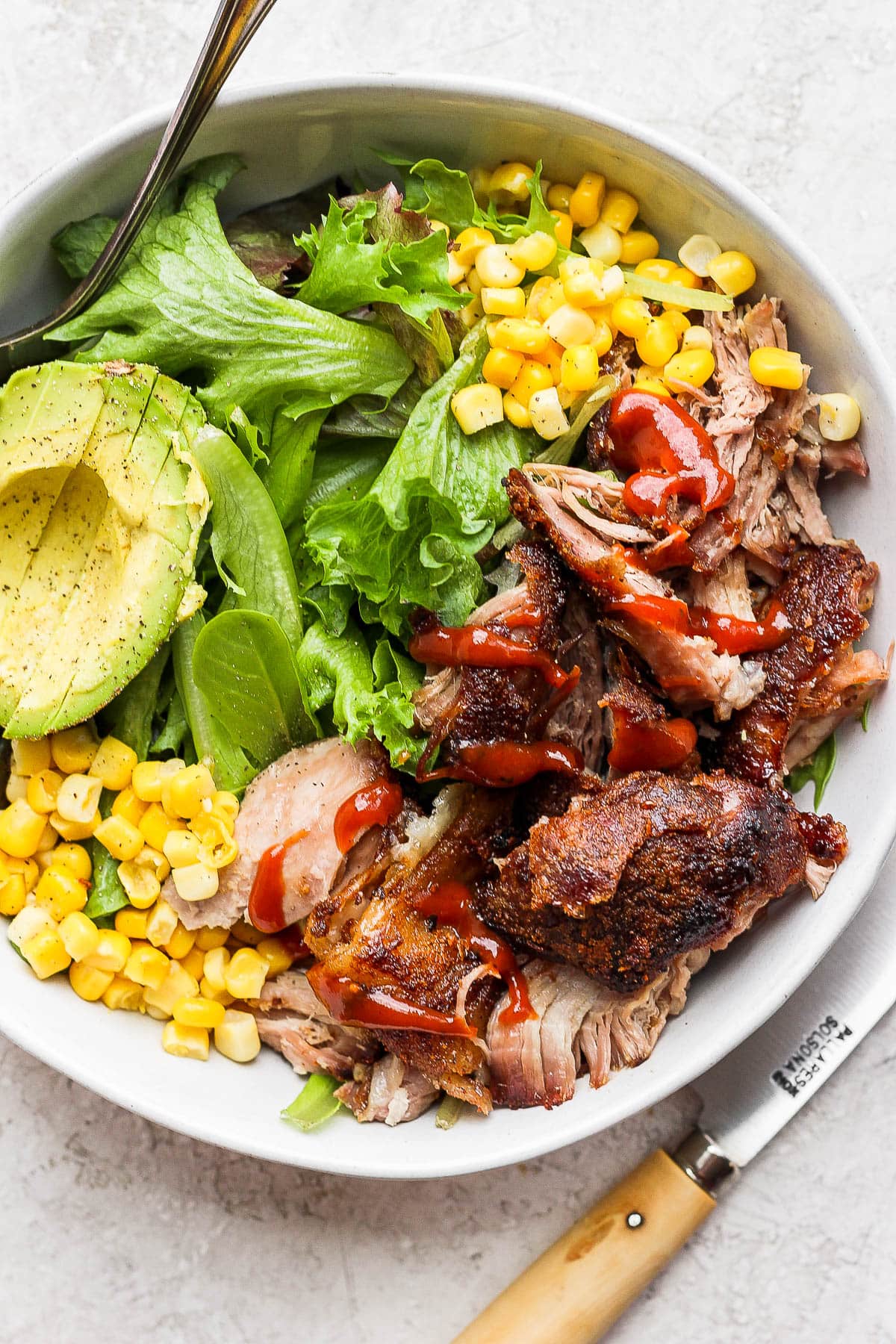 A visually appealing salad bowl is filled with mixed greens, sliced avocado, grilled corn, and pulled pork, all topped with a drizzle of red sauce, accompanied by a fork on the side.