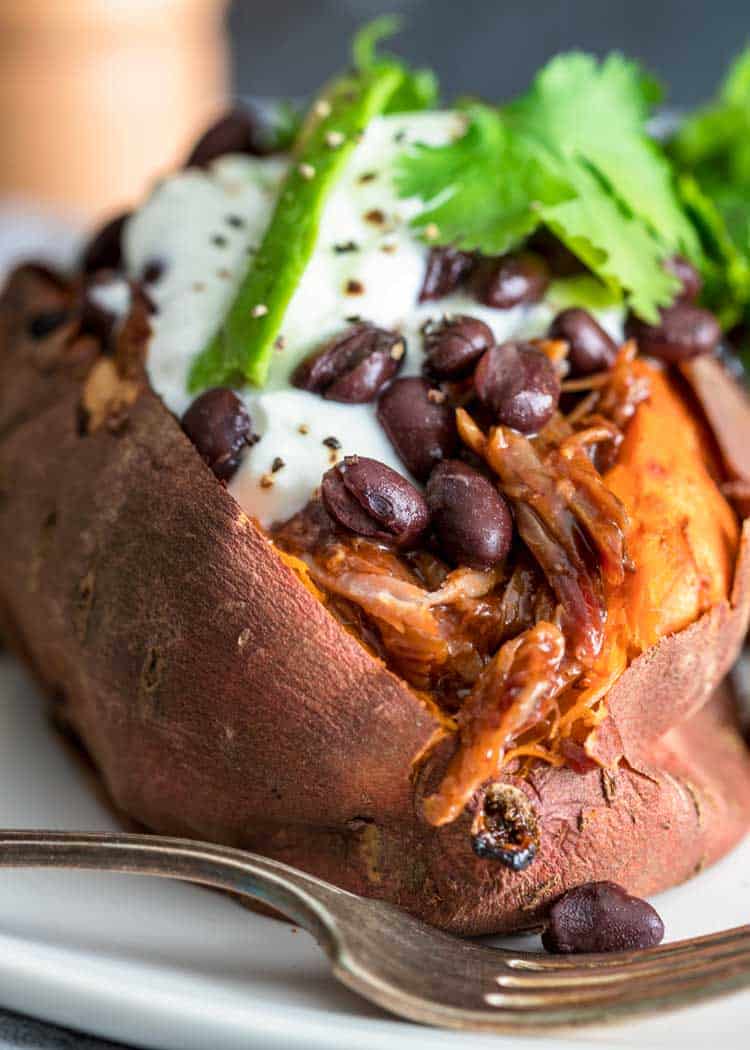 A sweet potato, baked and topped with shredded chicken, black beans, sour cream, and cilantro, is presented on a white plate accompanied by a silver fork.