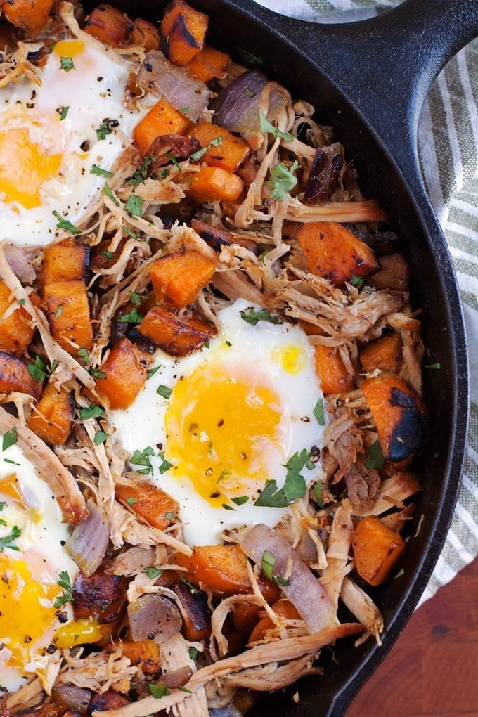 A skillet holds colorful sweet potato hash, shredded meat, sunny-side-up eggs on top, and is sprinkled with chopped herbs.