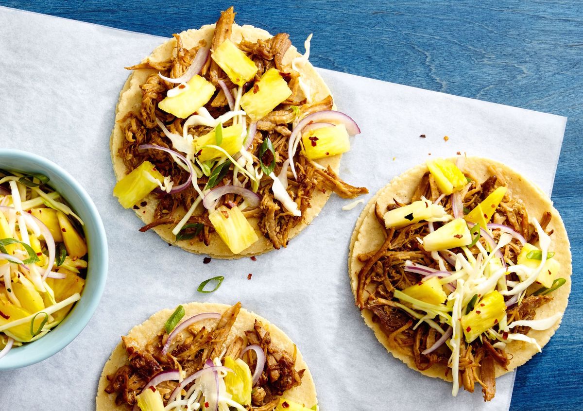 Three pulled pork tacos, garnished with pineapple, red onions, and cabbage, are arranged on a parchment-lined surface.