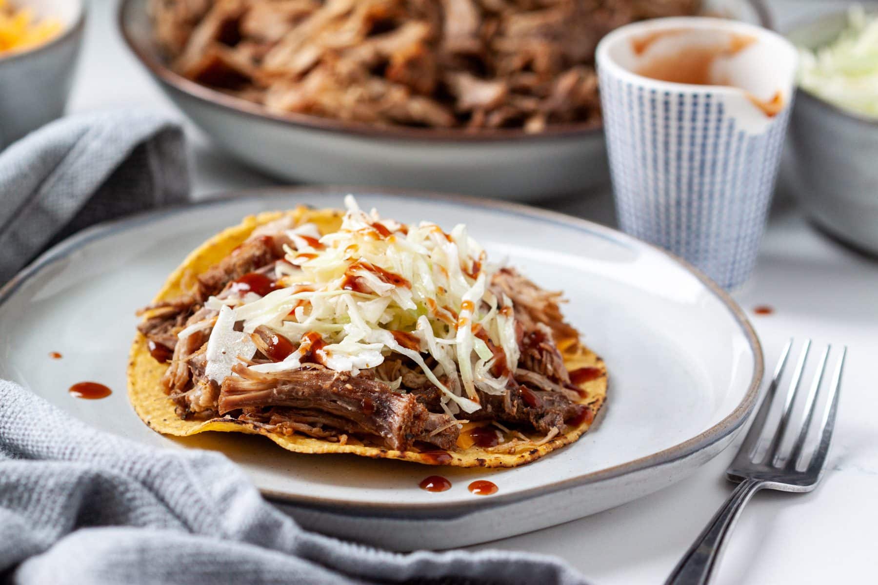 A plate displays a pulled pork taco garnished with shredded cabbage and sauce, alongside a side dish of extra sauce and a separate bowl filled with more pork in the background.