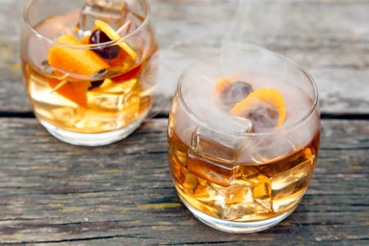 Two glasses of smoked old fashioned cocktails sit on a rustic wooden table with ice cubes, orange peels, cherries, and thin wisps of smoke rising from the drinks.