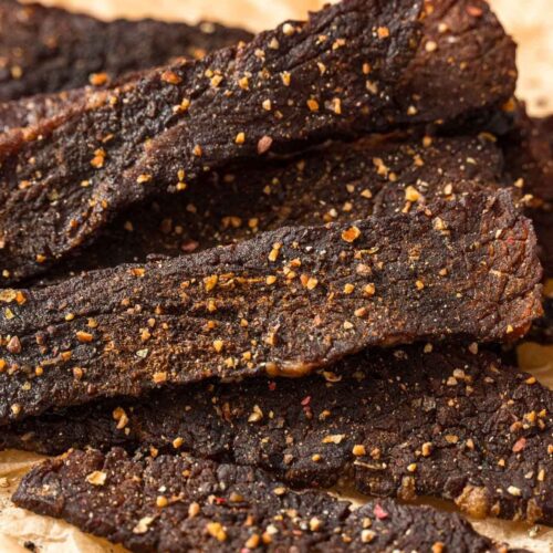 Close-up of several strips of dark, seasoned beef jerky on crumpled parchment paper, with visible spices and a textured, rustic appearance.