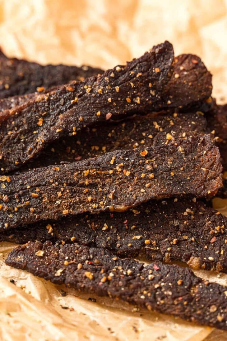 Close-up of several strips of dark, seasoned beef jerky on crumpled parchment paper, with visible spices and a textured, rustic appearance.