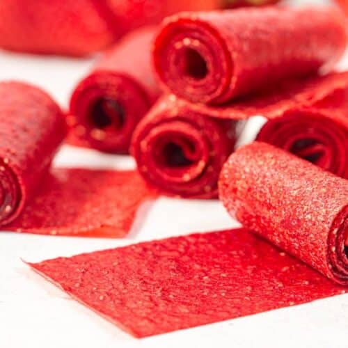 Several rolled and unrolled strips of red fruit leather are placed on a white surface, displaying their shiny, textured appearance.