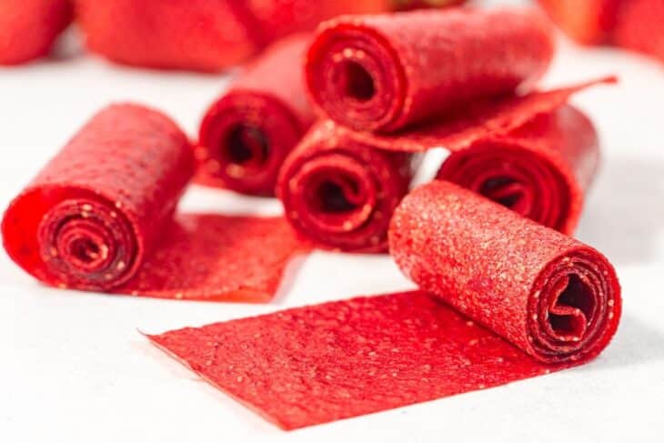 Several rolled and unrolled strips of red fruit leather are placed on a white surface, displaying their shiny, textured appearance.