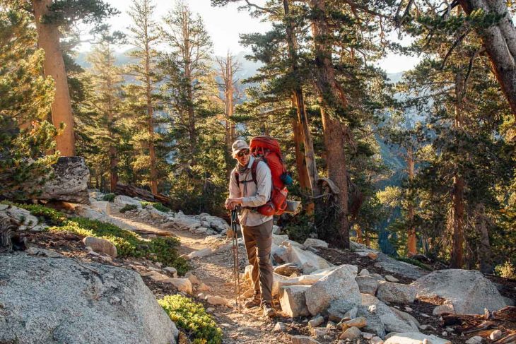 Michael is standing on the trail. He is wearing a long sleeve shirt and pants, a hiking backpack, and a hat and sunglasses