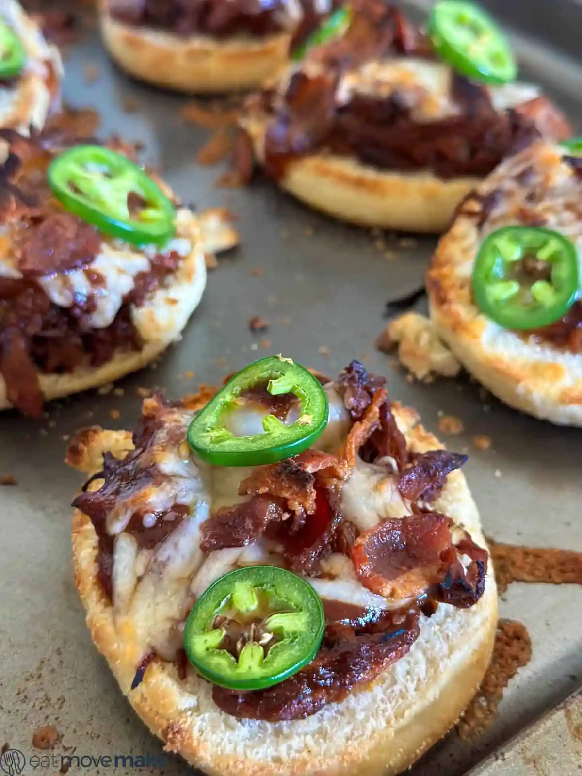 A tray holds mini pizzas garnished with strands of melted cheese, bits of crispy bacon, caramelized onions, and sliced jalapeños.