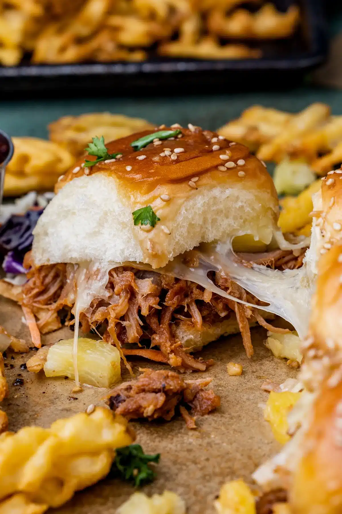 A pulled pork sandwich with melted cheese and pineapple on a sesame bun is seen close-up, accompanied by crinkle-cut fries in the background.