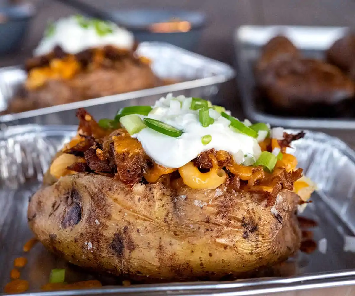 A baked potato in a small aluminum tray is garnished with shredded cheese, bacon bits, sour cream, and green onions.