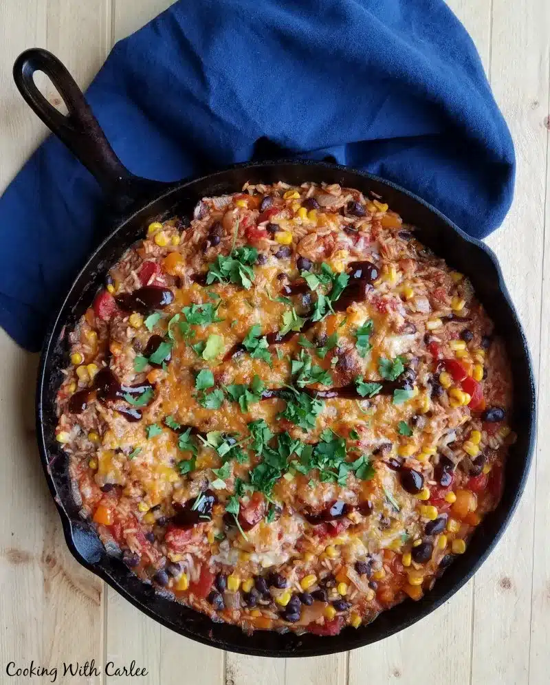 A skillet houses colorful mixed bean chili, accented with melted cheese and fresh cilantro, resting on a dark blue cloth background.