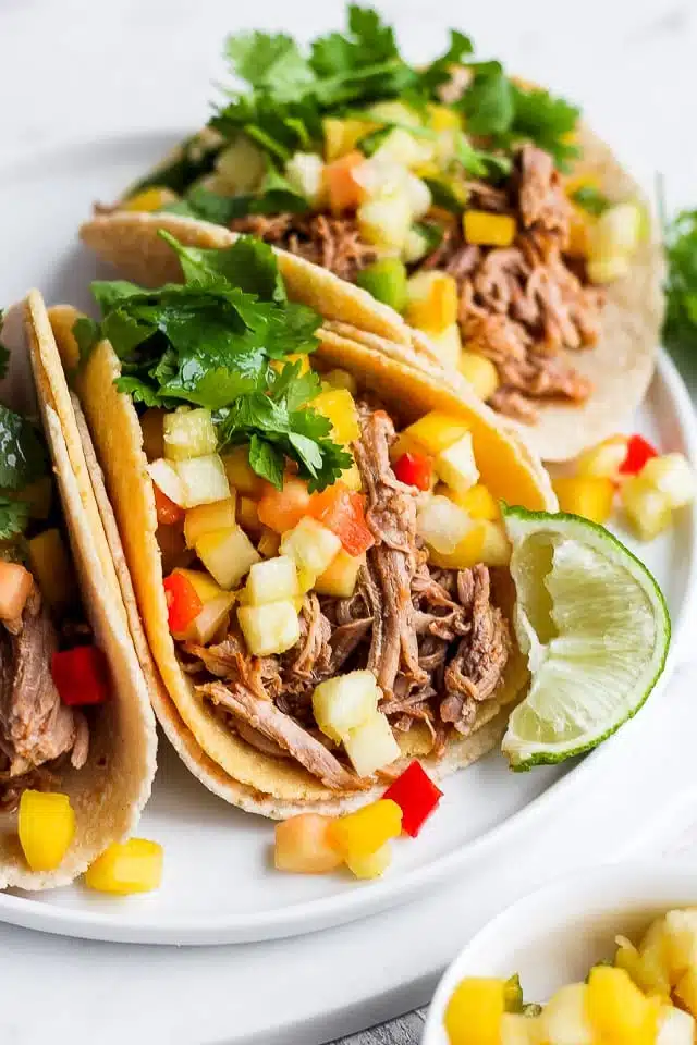 Three soft shell tacos, loaded with shredded meat, diced mango, red peppers, and cilantro, accompanied by a lime wedge on a white plate.