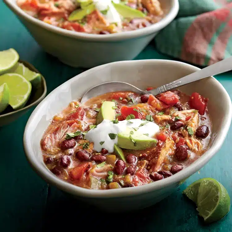 A bowl contains chili made of kidney beans, diced tomatoes, shredded chicken, topped with avocado slices and sour cream, accompanied by lime wedges.